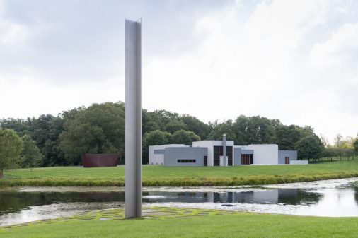 A tall, narrow, and silver metal sculpture stands at the edge of a pond. A gray, horizontal modernist building sits across the pond. 