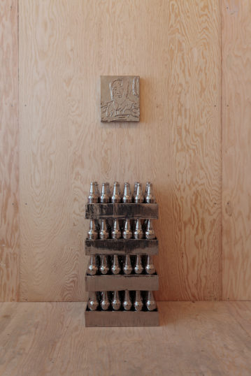 Four crates containing metallic beer bottles are stacked below a metallic portrait of Mao Zedong in a plywood room.
