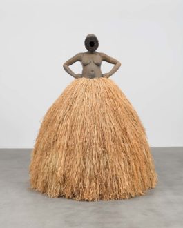 A sculpture of a woman with arms at her hips stands in a gallery space