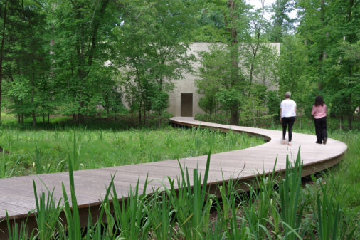Two figures approach a concrete rectangular building in the woods on a long and winding boardwalk.