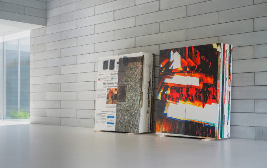 Two stacks of paintings sit in a room with a terrazo floor and concrete walls.
