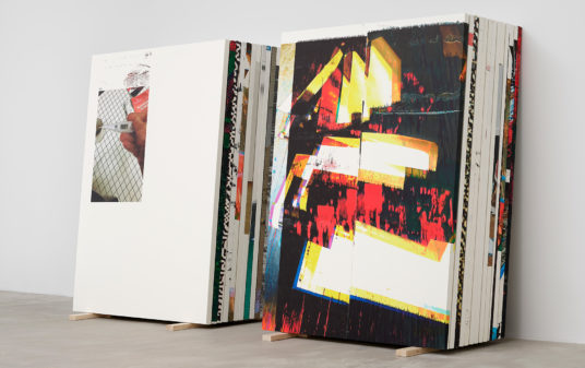 A stack of paintings sit on wooden struts on the floor of a gallery space.