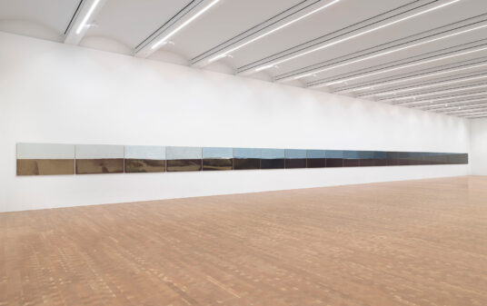A long painting, many panels wide, stretches across a white gallery wall.