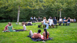 A crowd of people sit facing a jazz band on a green lawn.